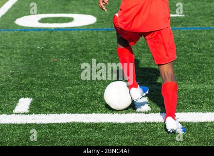 High School Fußballspieler trägt eine rote Uniform ist die Steuerung der Ball während eines Spiels auf einem grünen Rasen Feld. Stockfoto