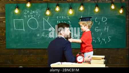 Junger männlicher Lehrer führt sein Kind Schüler zum Lernen während des Sprechens, sitzen im Klassenzimmer, Tafel mit Kritzeleien auf hinterem Hintergrund, Rückansicht Stockfoto