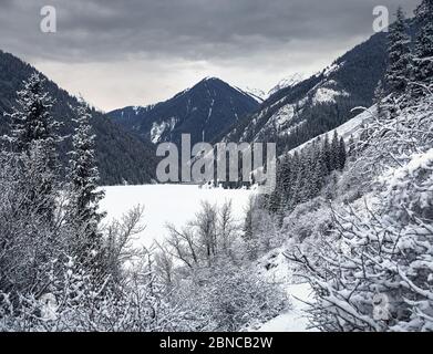 Landschaft von Kolsay-Bergsee und schneebedeckter Wald zur Winterzeit gegen bewölkten Himmel in den Bergen Kasachstans. Stockfoto