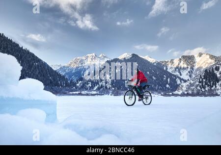 Der Mann in roter Jacke fährt mit seinem Fahrrad am gefrorenen See in verschneiten Bergen im Hintergrund Stockfoto
