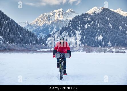 Das Porträt des bärtigen Mannes in roter Jacke ist ein Fahrrad am gefrorenen See in verschneiten Bergen im Hintergrund Stockfoto