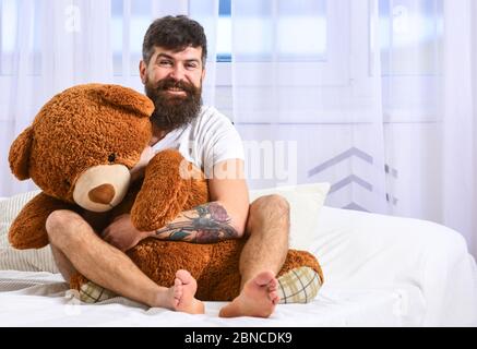 Kerl auf fröhlichem Gesicht umarmt riesigen Teddybär. Konzept des Infantilismus. Macho mit Bart und Schnurrbart Entspannen mit Plüsch Spielzeug nach dem Nickerchen, Ruhe. Mann sitzt auf dem Bett und umarmt großes Spielzeug, weiße Vorhänge auf dem Hintergrund. Stockfoto