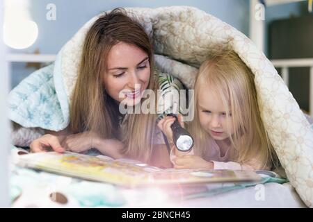 Junge Mutter liest ein Buch mit ihrem Kleinkind Mädchen mit Taschenlampe, während sie sich auf dem Bett Stockfoto