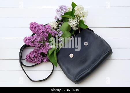 Frühling Lilac Blumen mit einer Leder-Handtasche in blau. Zweige der blühenden Flieder über einer hellen Holzoberfläche Frauen Tasche voller Blumen Draufsicht. Roman Stockfoto