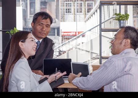 Drei asiatische Geschäftsleute und Geschäftsfrau diskutieren gemeinsam im modernen Bürogebäude mit Laptop und Tablet, Smartphone und Papierkarte. Stockfoto