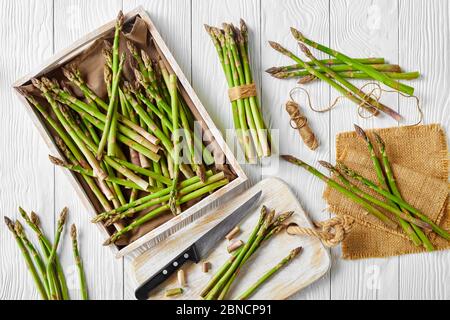 Bündel von frischem grünen Spargel auf einem rustikalen Holztablett und auf einem Schneidebrett mit Messer auf weißem Holztisch, horizontale Ansicht von oben, flaches Legen Stockfoto