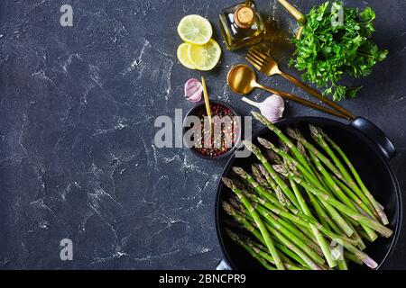 Frischer grüner Spargel in schwarzer Backform mit goldenem Besteck, Zitronenscheiben, Pfefferkörnern und einer Flasche Olivenöl auf grauem Betontisch, horizontal V Stockfoto