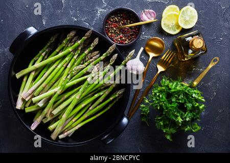 Nahaufnahme von frischem grünen Spargel in schwarzer Backform mit goldenem Besteck, Zitronenscheiben, Pfefferkörnern und einer Flasche Olivenöl auf grauem Betontisch, Stockfoto