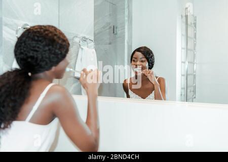 afroamerikanische Frau lächelnd, Zähneputzen und Blick auf Spiegel im Bad Stockfoto