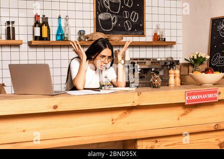 Schockiert Cafe Besitzer Blick auf Rechner in der Nähe von Papieren und Laptop am Tisch mit Karte mit Coronavirus Inschrift Stockfoto