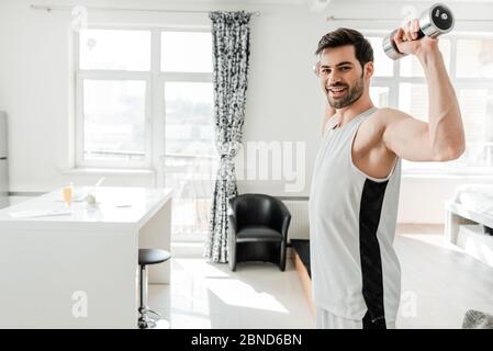 Seitenansicht des hübschen Mannes lächelnd auf Kamera beim Training mit Langhanteln zu Hause Stockfoto