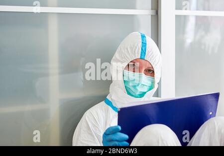 Pflegepersonal in der Klinik in Schutzkleidung mit Klemmbrett macht während der Coronavirus-Epidemie eine Pause auf dem Boden Stockfoto