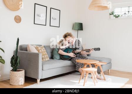 Tochter, die Mutter umarmt, während sie Gitarre spielt Zu Hause Stockfoto