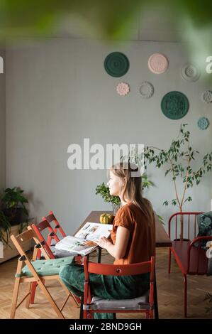 Frau sitzt am Tisch mit Zeitschrift, schaut auf Fenster im Wohnzimmer. Stockfoto