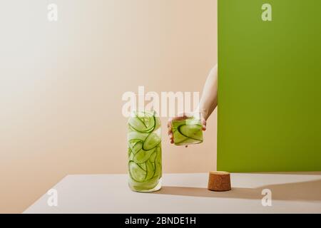 Cropped Ansicht der weiblichen Hand, die Glas mit Wasser gefüllt und Gurken in Scheiben auf beige und grün Hintergrund Stockfoto