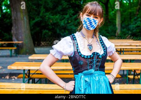 Frau in Dirndl, die im leeren Biergarten steht und eine blau-weiße Gesichtsmaske trägt Stockfoto