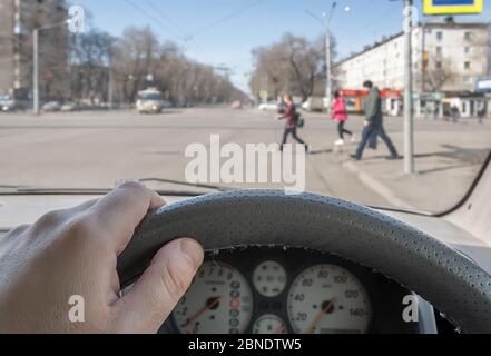 Blick aus dem Auto, Hand des Mannes am Lenkrad des Autos, befindet sich gegenüber der Fußgängerampel und Fußgänger über die Straße Stockfoto