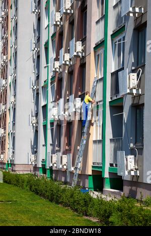 Ein Mann wäscht die Fenster auf der Straße, die auf der Treppe stehen. Reinigungsfirma. Arbeiter in blauen Overalls wäscht ein Fenster einer mehrstöckigen Wohnung Stockfoto