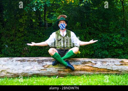 bayerischer Mann sitzt auf Baumstumpf und trägt eine blau-weiße Gesichtsmaske Stockfoto