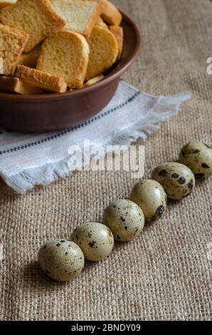 Stillleben mit Croutons und rohen Wachteleiern auf Burlap. Eine Tonschüssel mit kleinen quadratischen Semmelbröseln und einer Gruppe Wachteleier in einer Reihe. Einfach gesund Stockfoto