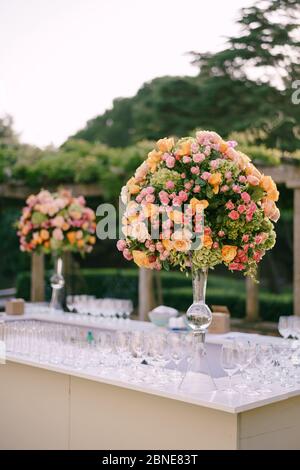 Empfang am Tisch für Hochzeitsessen. Riesige Blumensträuße aus gelben, rosa, orangen Rosen und grünen Hortensien in einer Glasvase auf einem hohen Bein. Große Blumensträuße in der Stockfoto
