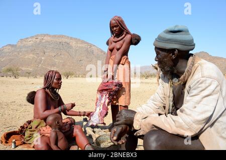 Himba-Mann beobachtet Frauen beim Fleischschneiden, Marienfluss Valley, Kaokoland Desert, Namibia. Oktober 2015 Stockfoto