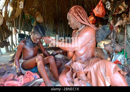 Himba Frau in ihrer Hütte, die Otjize (eine Mischung aus Butter, Ocker und Asche) auf die Haut ihrer Tochter aufsetzt. Marienfluss Tal. Kaokoland, Namibia Oktober 201 Stockfoto