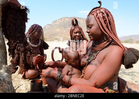 Himba Frau, die Otjize (eine Mischung aus Butter, Ocker und Asche) auf Röcke aus Ziegenhaut während des Stillens aufsetzt. Marienfluss Tal. Kaokoland, Nordamerika Stockfoto