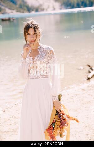 Schöne Braut in einem weißen Kleid mit Ärmeln und Spitze, mit einem gelben Herbststrauß von getrockneten Blumen und Pfingstrosen, am Lago di Prags in Italien Stockfoto