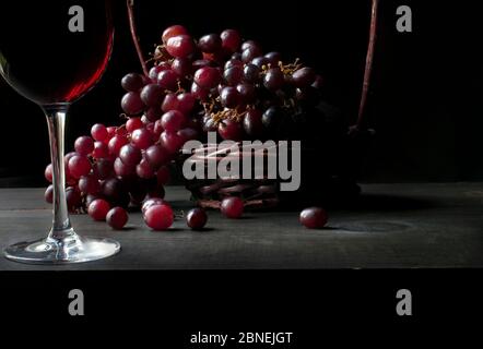 Stillleben, Glas mit Rotwein beleuchtet durch ein großes Fenster mit einem Korb mit roten Trauben auf einem schwarzen Holztisch und in einem schwarzen Hintergrund, Chiaroscuro, Stockfoto