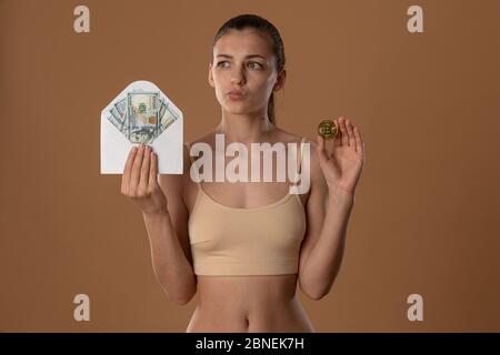 Discomposed hübsches Brünette Mädchen mit natürlichen Make-up in beige BH gekleidet hält weißen Umschlag in einer Hand und Bitcoin goldene Münze in anderen Stockfoto