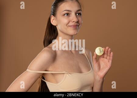 Zufrieden Brünette Mädchen mit natürlichen gesunden Haut hält Bitcoin goldene Münze in der Hand und Blick auf die rechte Seite Stockfoto