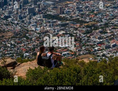 Kapstadt, Südafrika - 17. März 2020:Junges Paar auf dem Felsen auf dem Gipfel des Lion's Head Mountain sitzend, schaut auf das Stadtbild von Kapstadt. Stockfoto