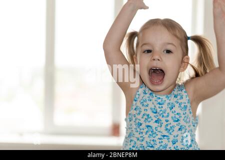 Kindheit, Menschen, Emotionen Konzept - mittelgroßer Plan kleines Mädchen mit zwei Pferdeschwänzen und einem blauen Kleid springt die Hände hoch und schreit am Fenster. Stockfoto