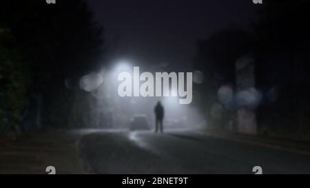 Eine geheimnisvolle Figur, die in einer nebligen Winternacht mitten auf einer Straße mit Straßenlaternen steht. Mit einem unscharfen, Bokeh, unscharf bearbeitenden Schnitt Stockfoto