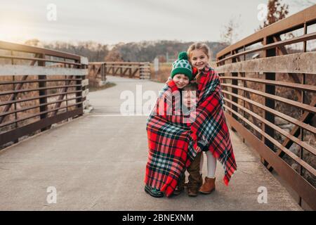 Geschwister, eingewickelt in eine karierte weihnachtsdecke auf der Brücke bei Sonnenuntergang Stockfoto