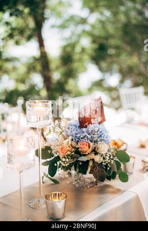 Empfang am Tisch für Hochzeitsessen. Blumenstrauß in einer Vase auf einem Tisch aus rosa und weißen Rosen und blauen Hortensien, mit der Nummer des Tisches auf dem Teller Stockfoto