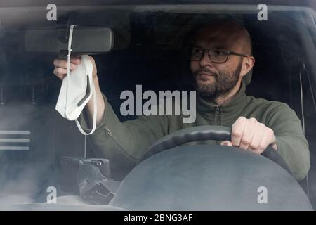 Porträt des jungen Mannes im Auto sitzen, die für schützende Gesichtsmaske, die vom Rückspiegel im Auto hängt, von vorne gesehen, durch vorne gesehen, Shi Stockfoto