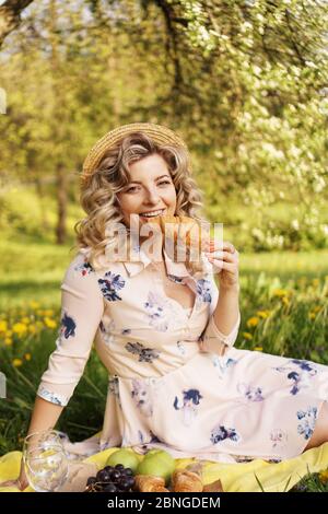 Schöne junge Frau mit blonden Haaren in Strohhut und weißem Kleid auf dem Plaid im Park liegen. Frau mit einem Croissant auf einem Picknick Stockfoto