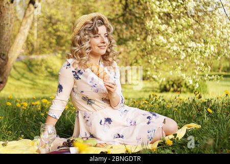 Schöne junge Frau mit blonden Haaren in Strohhut und weißem Kleid auf dem Plaid im Park liegen. Frau mit einem Croissant auf einem Picknick Stockfoto