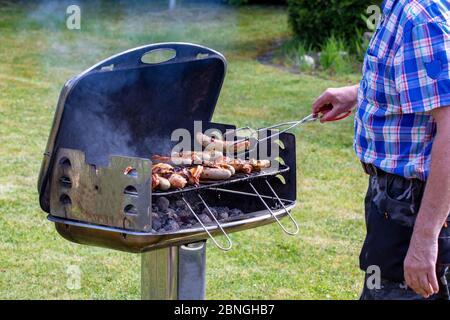 Ein Mann am Holzkohlegrill grillen Steaks, Grillwürste und Hühnerflügel und benutzen Grillzangen. Selektiver Fokus auf den Grill gegen eine gree Stockfoto