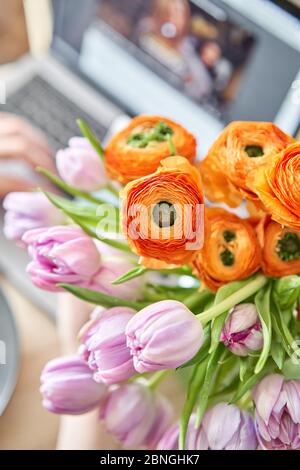 Frühlingskomposition mit lila Tulpen und orangefarbener Butterblume und einer Tasse grünen Tee mit Zitrone, einem offenen Laptop auf einem Holztisch. Konzept für den Aufenthalt zu Hause Stockfoto