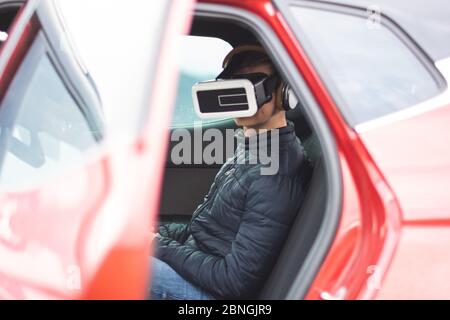 Blick auf einen Mann, der die virtuelle Realität benutzt, während er im Sitzen sitzt Der Rücksitz eines roten Autos mit seiner Tür Öffnen Stockfoto