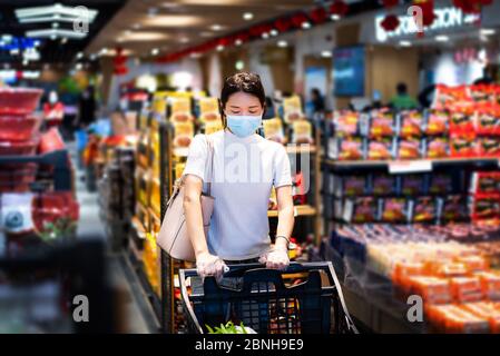 Asiatische Frau, die auf dem Markt Lebensmittel kauft, trägt Maske und schiebt den Wagen mit Handschuhen, um die Ausbreitung des Virus während der Coronavirus-Pandemie zu verhindern Stockfoto