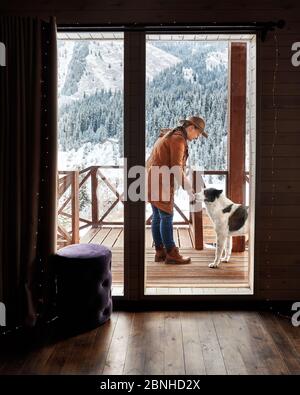 Frau in Hut spielt mit ihrem Hund im Winter im Holzhaus im Bergresort Stockfoto