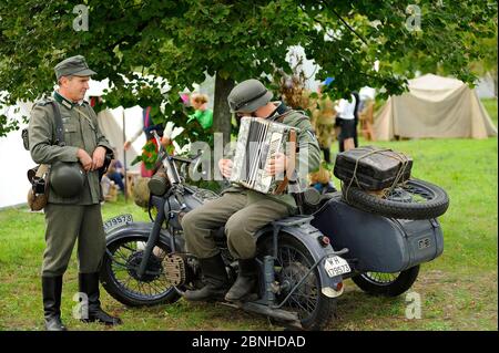 Reenactor in Uniform des deutschen Soldaten des Zweiten Weltkriegs auf einem alten Militärmotorrad sitzend und Akkordeon spielend gekleidet Stockfoto