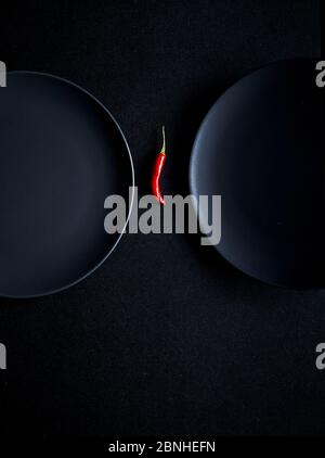 Minimalistische Komposition aus rotem Chilli zwischen zwei Keramikplatten auf schwarzem Hintergrund Stockfoto
