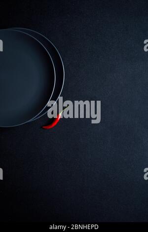Minimalistische Komposition aus rotem Chilli in der Nähe von zwei Keramikplatten auf schwarzem Tisch Stockfoto