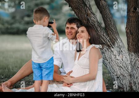 Glückliche Familie entspannt in der Natur unter einem Baum am Nachmittag. Das Kind fotografiert seinen Vater und seine schwangere Mutter Stockfoto
