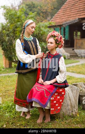 Zwei schöne Mädchen in Nationalkleidung des späten 19. Jahrhunderts. Eine Frau webt Bänder in das Haar ihrer Tochter. Alte Kleidung. Retro-Inszenierung eines Stockfoto